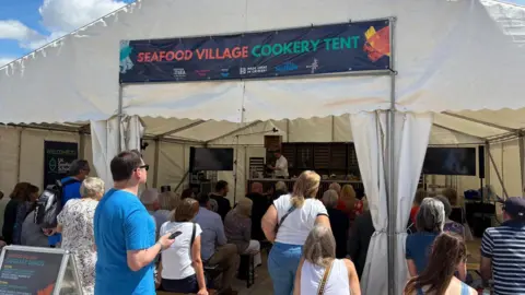 North East Lincolnshire Council A large white tent with a blue sign on reading "SEAFOOD VILLAGE COOKERY TENT" in colourful capital letters. People are gathering, either sat or stood, inside and outside of the tent. 