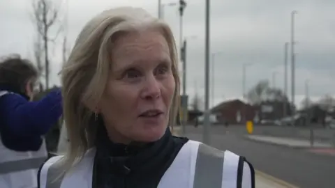 Janine Bowyer, who is wearing a Crosby Wombles safety vest, is standing on a path on the side of a road during a litter picking session. 