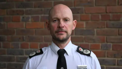 Jason Hogg, dressed in police uniform, stood in front of a brick wall.