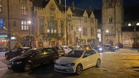 Several cars parked in the centre surrounded by gothic medieval-looking buildings at night-time.