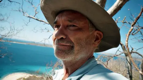 Channel 5 Phillip Schofield smiling in front of a beach and the sea