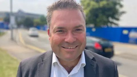 Michael Whitehouse smiling and wearing grey suit standing in front of road.