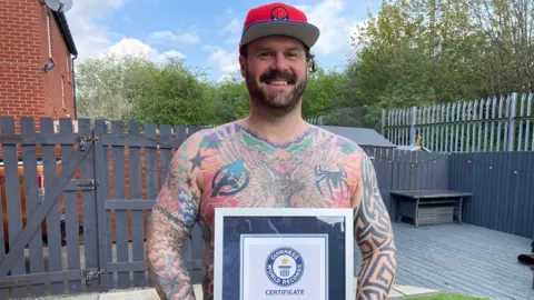 Tom Radford stands bare chested showing his tattoos. He has a black beard and moustache and is wearing a red Spiderman baseball cap