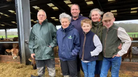 BBC / Peter Whittlesea Six members of the same family - two men, two women and two boys - stand in front of a cow shed. 