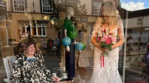 Alec W G Hall Ltd Val wears a black and white flowery outfit with a hat of black net. Aimee is in bridal wear and has borrowed the flower bouquet seen in the other picutre. Her dress is made up of chiffon and lace.