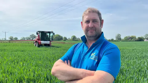 BBC Photo of Lay looking into the camera with his arms crossed. He's wearing a bright blue polo shirt with the collar turned up. He's stood in a wheat field, with a red combine harvester behind him. The sky is blue and there is lots of sun. 