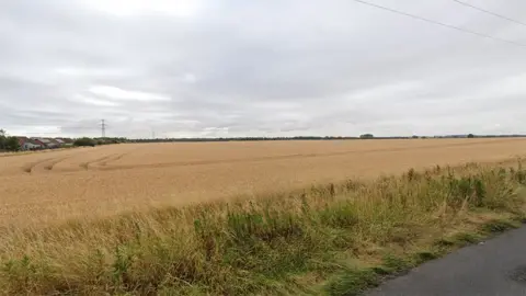 Google street view of part of the proposed site showing a field of crops with housing in the distance and electricity pylons overhead