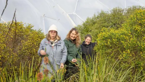 Three women standing in a line next to greenery. Behind them is the Eden Project's while biome. 
