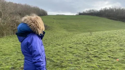 A woman in a thick blue coat stands looking up at a large grass hill.