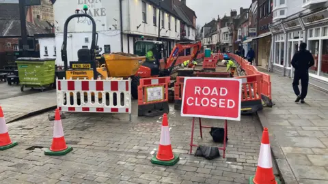 Roadworks in a cobbled street. Four red and white cones, red and white barriers and a large red "Road Closed" sign block the way. Behind the barriers, workers in hi-vis clothing can be seen working on the road, with one operating a small digger. A whitewashed pub and rows of traditional-looking shops line the route. Pedestrians can be seen walking down the pavement, which remains open.