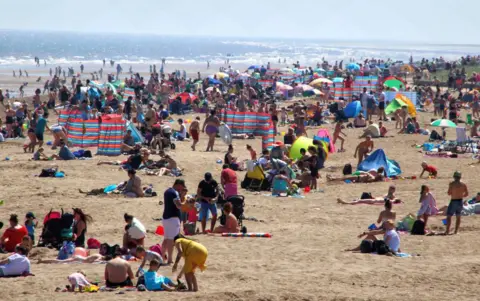 SOPA Images/Getty A crowded sandy beach packed with groups of people, colourful windbreaks and umbrellas. The sea and waves are visible in the background on a sunny day.