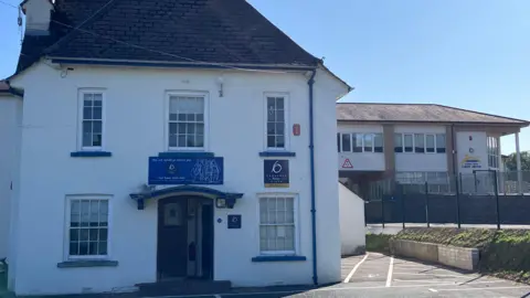 The entrance of Ysgol Bro Caerinion with a door leading to a white school house. There is a road to the right that runs underneath a second floor bridge and a school yard can be seen on the right hand side. 