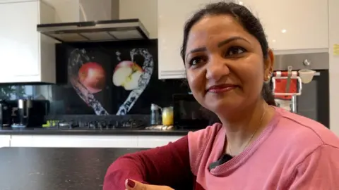 Sudha Sri Nukana sits at her kitchen table and faces the camera. She wears a pink jumper with darker pink sleeves and red lipstick.