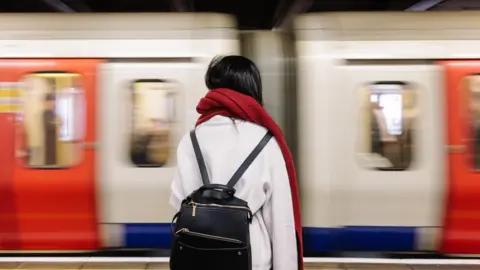 A file image of a woman with backpack and red scarf waiting on a platform as a Tube train goes past. 