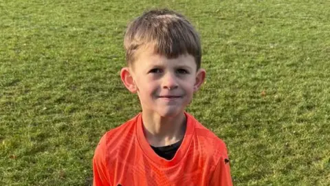 British Heart Foundation Archie wearing an orange football kit while standing on pitch. He is smiling at the camera.