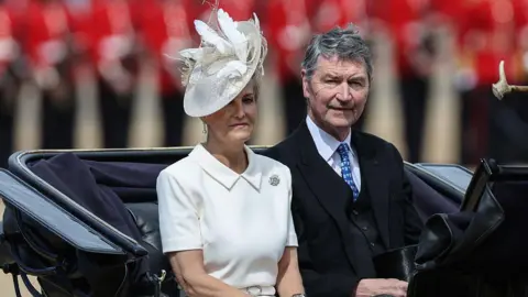 Getty Images The Duchess of Edinburgh and Vice-Admiral Sir Tim Laurence ride in a black carriage. She is wearing a white dress with a collar and a brooch with a white hat. He is wearing a dark suit with a blue tie.