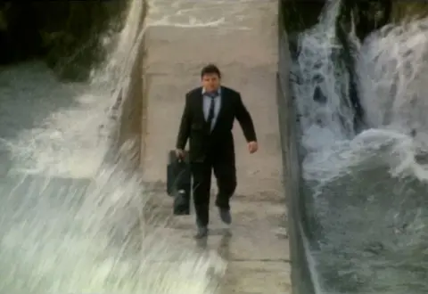 The Supergrass (1985)/A Peter Richardson Film Actor Robbie Coltrane, wearing a suit and holding a guitar case, walking along the Hope Cove Harbour breakwater in strong seas and high waves in the film The Supergrass (1985).