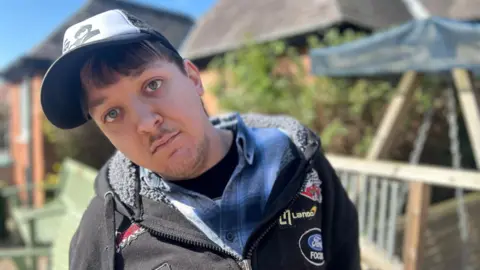 A young man in a baseball cap with black hooded jacket and blue shirt sat in a garden