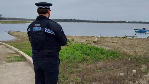 Essex Police A uniformed policeman stands with his back in view. He is facing a large expanse of water making up the reservoir. 