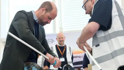 PA Media Prince William holding a long thin white pipe in his right hand and cutting it with a small blue pipe cutter held in his left hand. He is being watched by two men wearing white hi-viz vests.