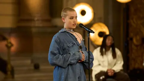 ITV Ace, wearing a long oversized denim shirt and talking into the mic during the callback round of the voice. She has short blonde hair and behind her are other contestants sat down