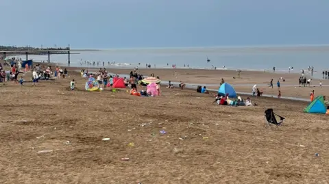 RNLI Lifeguards Lincolnshire Sandy beach with litter on it. There are people and tents and a folding chair. There are pylons in the distance There is a sea.