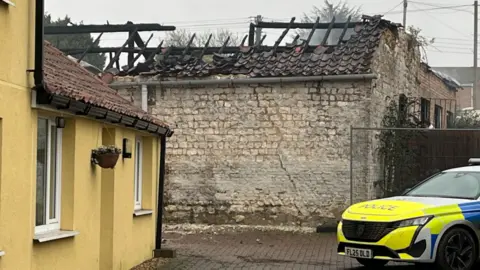 A stone barn with the burnt-out remains of a roof. On the right is the front of a police car and on the left is a yellow-painted house.