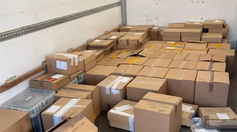 City of London Police Dozens of brown cardboard boxes in rows and stacked on top of one another. The boxes are secured with brown and white tape.