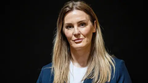 PA Media UUP councillor Linzi McLaren has blonde hair with brown roots. She is standing in front of a black background wearing a blue blazer and a white t-shirt, smiling.