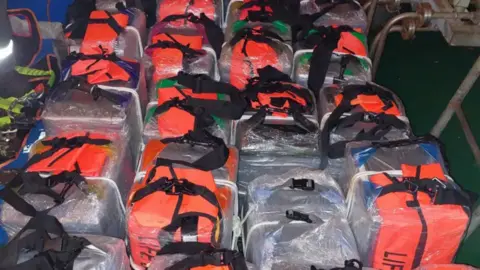 Border Force A shot of about 20 orange and black bags lined up on the ground. The bags are wrapped in plastic