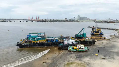 An aerial view of the city of Toamasina, on the east coast of Madagascar. Three boats are grounded on a sandy shoreline beside a wide bay. Machinery and equipment sit on the vessels, with one tugboat tilted at an angle on the beach. A person walks nearby, and industrial buildings and cranes are visible across the water in the distance.