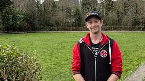 Simpa Carter smiling into the camera while standing in front of a square patch of cut grass. He has brown stubble around his mouth and is wearing a black cap and red and black zip-up up hoodie and black trousers. He has his hands clasped in front of his body. Carter is standing on a path to the right of the grass and in between two hedges which run all the way round the land. There are trees beyond the green, most of which are without leaves.