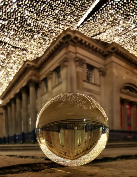 Jane Reith Evening image of the Gallery of Modern Art in Glasgow, taken from street level with sheets of fairy lights hung above Royal Exchange Square. A decorative glass ball is in the foreground and the museum is inverted in the reflection of the ball.