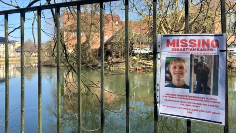 A missing poster for Sebastian, on white paper with red lettering, attached to a railing next to a large body of water. 