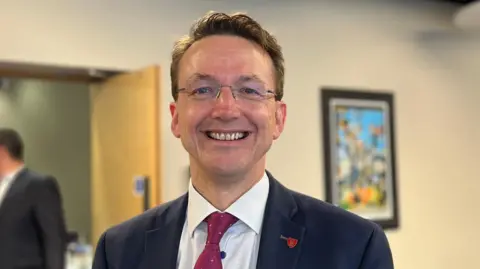 Deputy Kirsten Morel, a man with short brown hair and glasses with no frame. He is staring directly at the camera and smiling. He is wearing a white shirt, maroon tie and a navy blue blazer. He has a red lapel badge. Behind him, out of focus, a colourful piece of artwork is visible on the wall, in a dark frame.