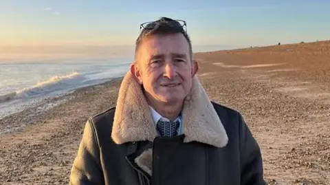 Councillor David Wimble stands on a beach in a leather jacket with a woolly collar