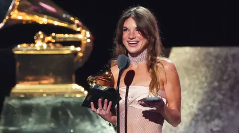 Amy Allen on stage at the Grammy Awards, standing in front of a microphone, holding a Grammy in her right hand and a mobile phone in her left - she has long wavy dark hair and is wearing a strapless pale pink satin and mesh dress, and is smiling 