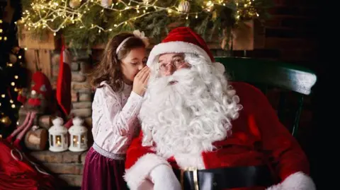 Getty Images A young girl visits Santa and whispers into his ear. There are fairy lights and Christmas decorations behind them and red ribbons.