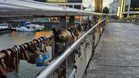 A metal footbridge over a harbour. The bridge is covered in padlocks. Boats are visible on the water and the lights of bars and restaurants can be seen in the background. 