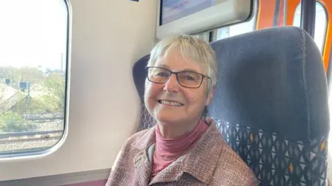 An elderly woman with short white hair sits on a train while wearing a pink jacket and pink jumper.