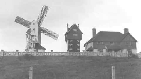 Charlotte de Mille/Ogilvie family A black and white image of a windmill, a large tower with a house on the top and a country club house at the top of a hill.