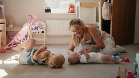 Family handout Becki is crouched down over her baby boy in a play room in there house. Her little girl is lying on the floor opposite. There are toys surrounding them. 