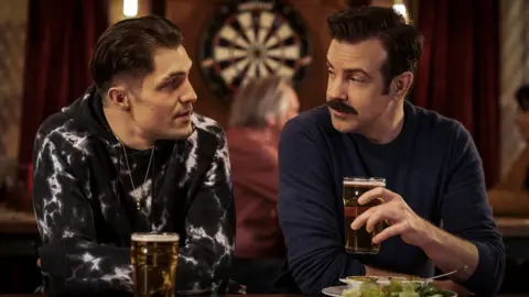 Apple TV+ Actor Phil Dunster as his character Jamie Tartt sat in a pub with Jason Sudeikis as his character Ted Lasso. Jamie has a pint of beer in front of him and Ted is holding a pint of beer in his left hand. There's a dartboard in the background behind them