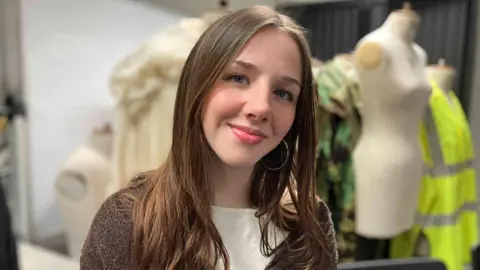 Alex Dunlop/BBC Scarlett Baker has long brown hair and is wearing a textured brown cardigan over a light top. Behind her, there are some mannequins, one features a cream‑coloured dress, another has a green patterned outfit, and a third is dressed in a bright yellow high‑visibility jacket but they are not in focus. 