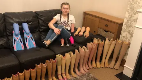 Olivia Story as a child sitting on a black sofa and smiling with her legs up. On the floor in front of her are a series of prosthetic legs lined up. There is a chest of drawers in the corner of the photo. Story has ponytails and brown hair. 