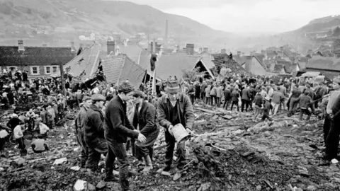 Getty Images A black and white image showing a mound of coal waste smothering the ruined Pantglas Junior School in Aberfan. Dozens of rescue workers can be seen standing near the roof of the school, which is surrounded by slurry. Some men in the foreground are emptying buckets of waste which are being passed back along a human chain. In the background are rows of terraced houses and a chimney stack, along with the hillside on the opposite side of the valley.