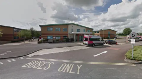 A school building in front of a car park. Words written on the road say buses only, and an arrow points  for the direction of traffic.
