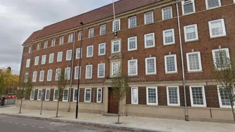 Google The large brick building with rows of windows has a front door in the centre and a pavement outside with a few small trees in front.