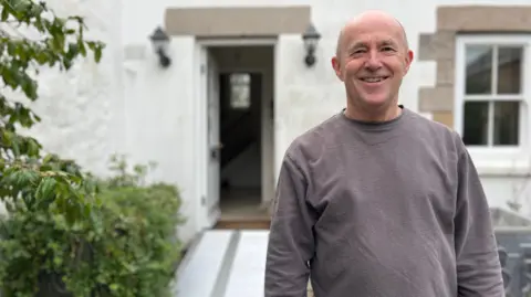 Nick is standing outside in front of a white building with an open door. A metal accessibility ramp leads into the doorway, and there are plants on the left side of the image.