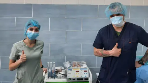 A male and female doctor hold their thumbs up in a hospital, next to some medical equipment. They wear medical caps, masks and scrubs.
